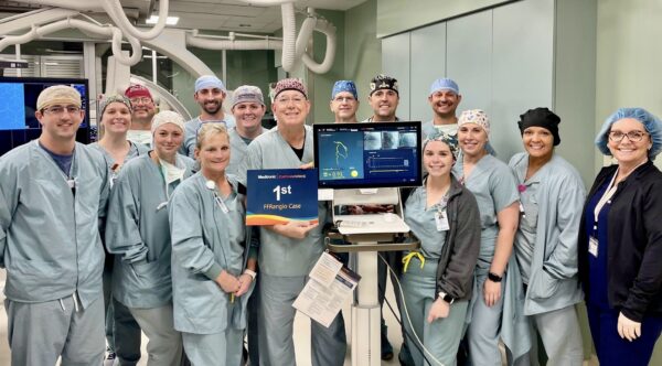 A group of medical staff in scrubs, including surgeons and nurses, posing in an operating room with medical equipment and screens displaying surgical data.