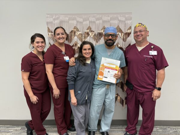 Five healthcare workers pose together indoors, one holding a certificate. Four wear maroon scrubs, one wears blue scrubs and a surgical cap.