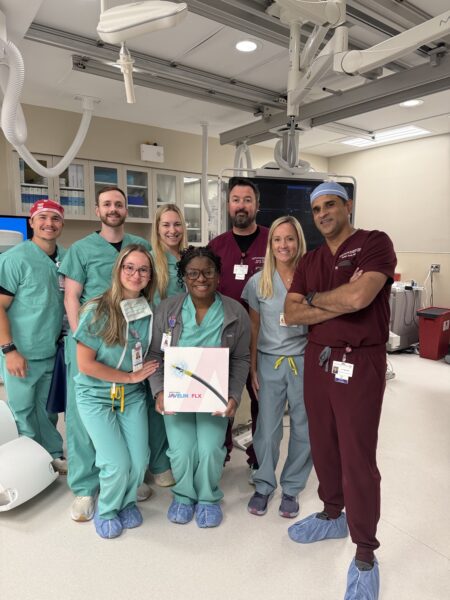 A group of eight medical professionals pose together in a hospital room, with one person holding a box labeled "AMPLATZER AMULET.