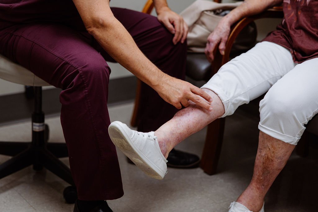 A healthcare worker examines the lower leg of a seated woman wearing white pants and white shoes in a clinical setting.