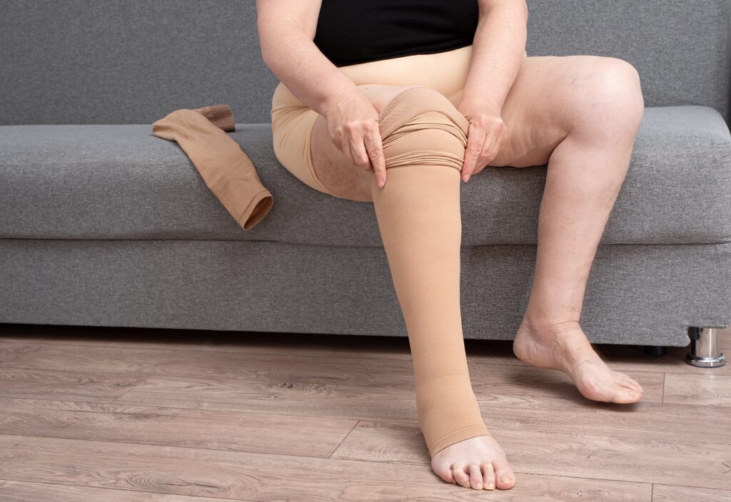 A woman sitting on a couch pulling on a beige compression stocking on her right leg.