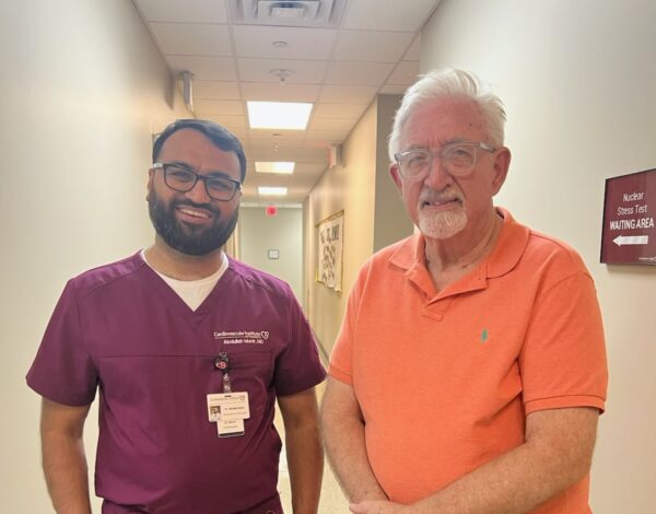 A man in purple medical scrubs stands next to an older man in an orange polo shirt and white shorts in a hallway.