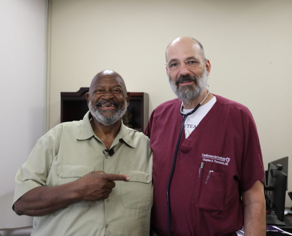 Two men stand together in an office; one is wearing medical scrubs with a stethoscope, and the other is pointing at him and smiling.