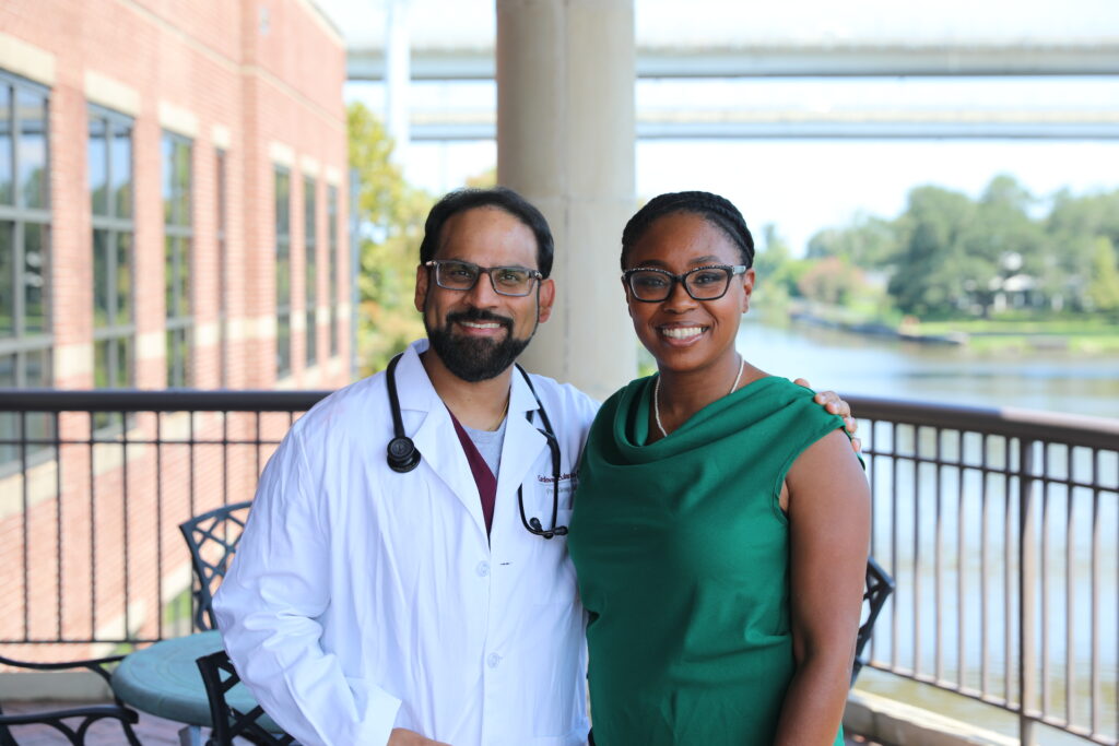 A man in a white coat with a stethoscope stands next to a woman in a green dress, both smiling, outdoors by a river.