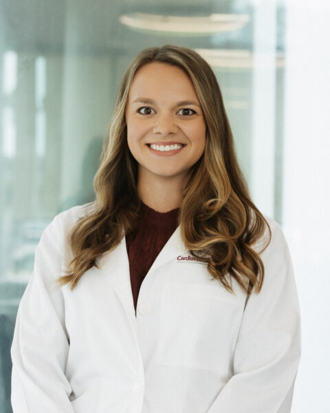 A woman with long brown hair wearing a white lab coat and maroon top stands smiling in front of a glass window.