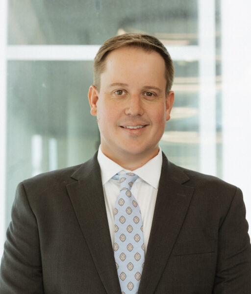 A man in a dark suit, white shirt, and patterned tie stands indoors in front of a window with a neutral expression.
