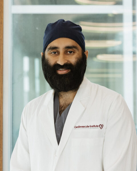 A man wearing a white lab coat and dark surgical cap stands indoors near a glass wall, looking at the camera and smiling.