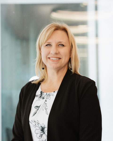 A woman with blonde hair, wearing a black blazer and floral top, stands indoors and smiles at the camera.