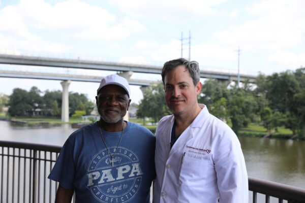 Two men stand side by side outdoors by a railing, with water, greenery, and bridges visible in the background.