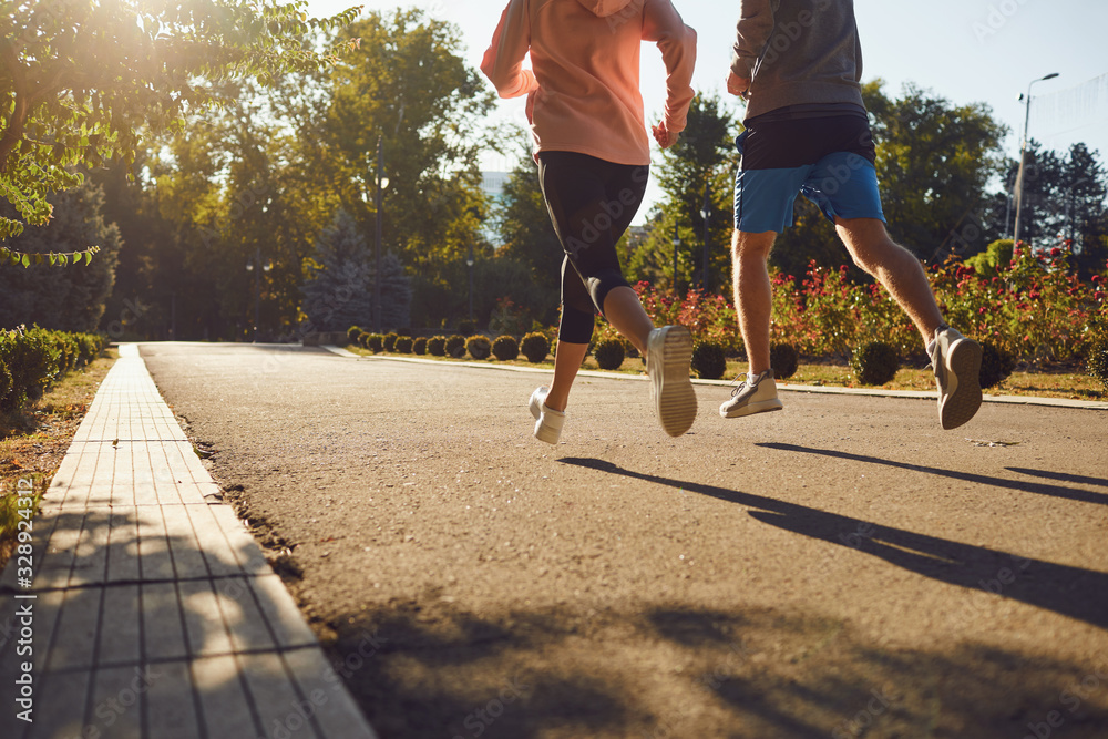 Two people are jogging on a paved path in a park on a sunny day, surrounded by trees and greenery.
