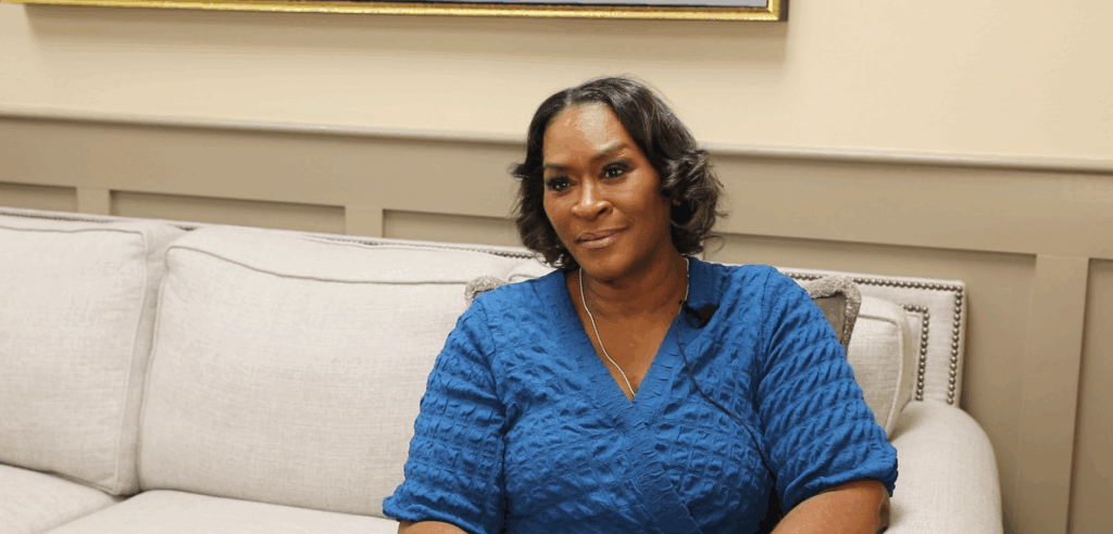 A woman in a blue textured top sits on a light-colored couch, looking slightly to the side in a softly lit room.