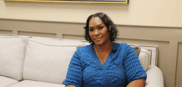 A woman in a blue textured top sits on a light-colored couch, looking slightly to the side in a softly lit room.