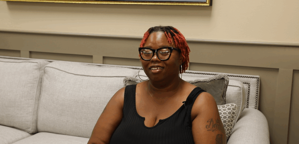 A woman with red hair and glasses sits on a light gray couch, wearing a black sleeveless top, smiling slightly at the camera.
