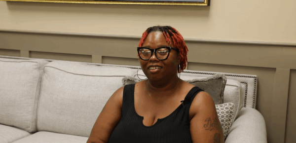A woman with red hair and glasses sits on a light gray couch, wearing a black sleeveless top, smiling slightly at the camera.