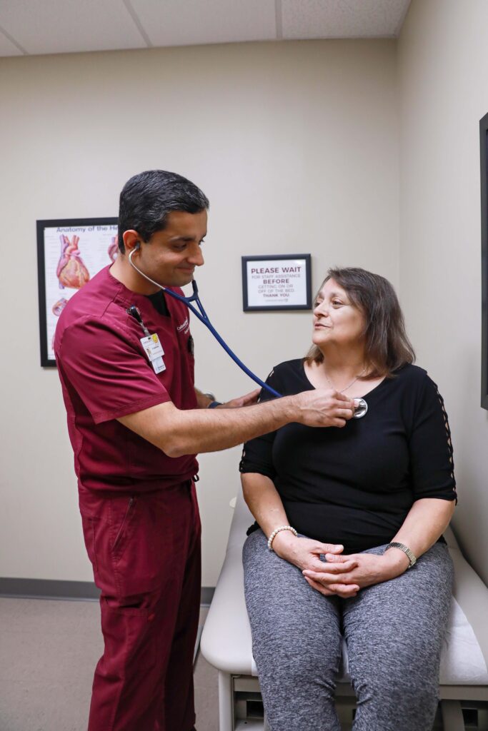A healthcare professional uses a stethoscope to check a seated patient's chest in a medical examination room.