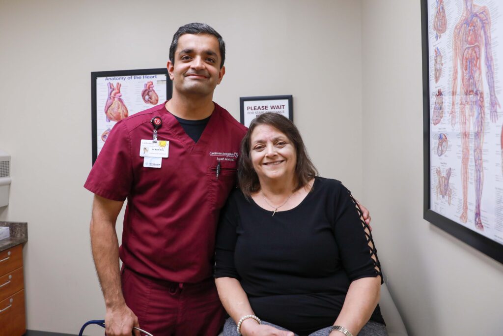 A healthcare professional in maroon scrubs stands next to a seated woman in a medical exam room, both facing the camera and smiling.