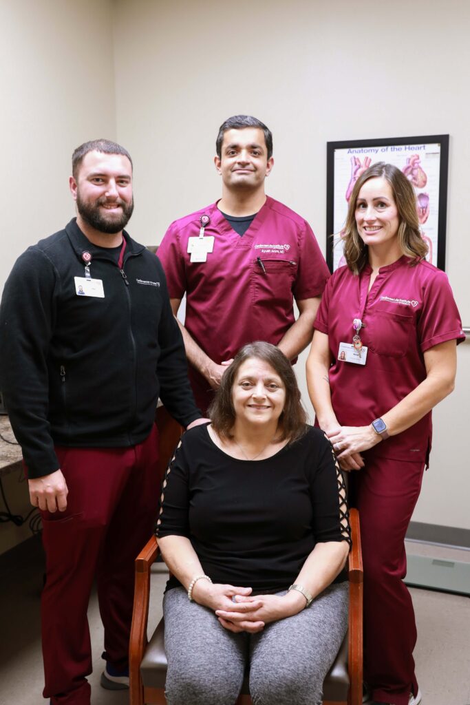 Four medical professionals pose with a seated woman in a clinic room, with a medical poster on the wall behind them.