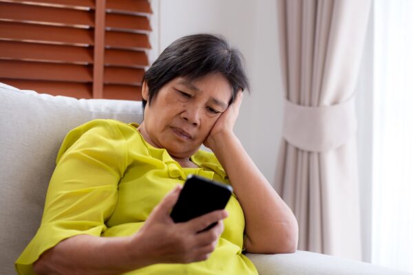 Older woman in a yellow shirt sitting on a couch, looking at her phone with a serious expression
