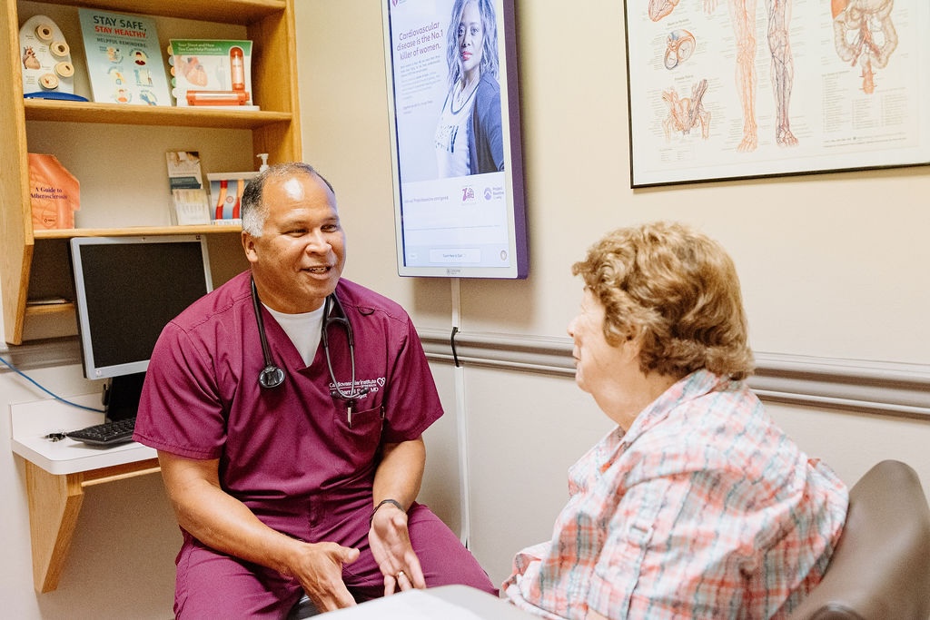 A doctor in maroon scrubs talking with a seated patient in a medical office