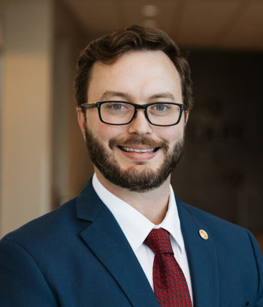 A man wearing glasses, a blue suit, white shirt, and red tie, standing indoors and smiling at the camera.
