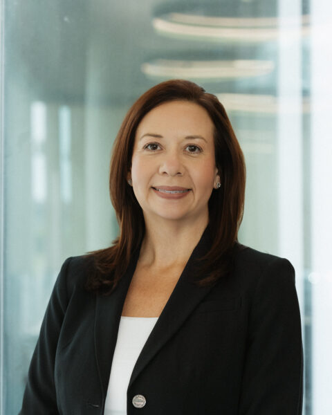 Woman with straight brown hair wearing a black blazer and white top, standing in an office with glass walls and ceiling lights.