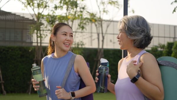 Two women in athletic wear holding water bottles and yoga mats while smiling and talking outdoors on a sunny day.