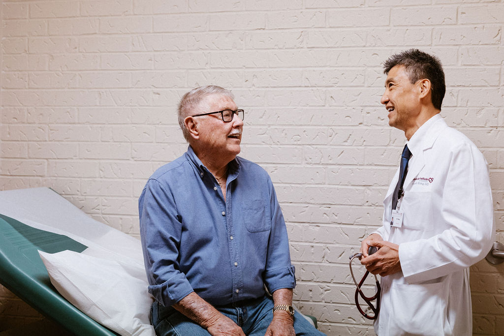 An older man sits on an exam table, talking with a doctor in a white coat holding a stethoscope in a medical exam room.