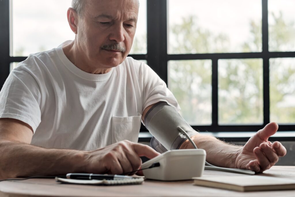 An older man checks his blood pressure using a digital monitor at a table near a window.