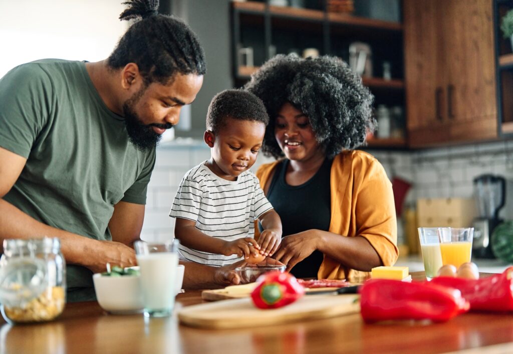 A man, a woman, and a young boy prepare food together in a kitchen with vegetables and drinks on the counter.