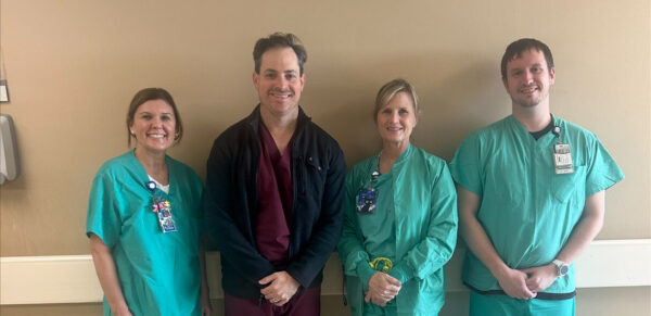 Four healthcare professionals in scrubs stand side by side against a beige wall, smiling at the camera in a hospital hallway.