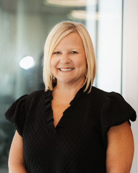 A woman with straight blonde hair wearing a black textured blouse stands indoors, smiling at the camera.