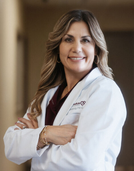 A woman wearing a white lab coat stands indoors with her arms crossed, smiling at the camera.