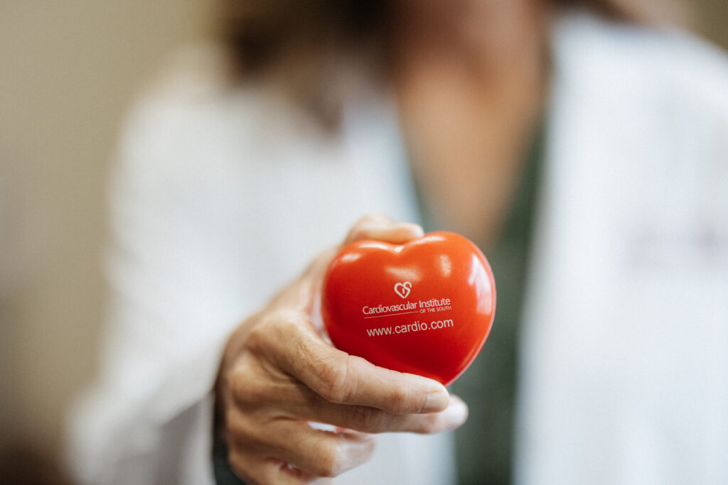 A person in a white coat holds a red heart-shaped stress ball with "Cardiovascular Institute" and a website printed on it.