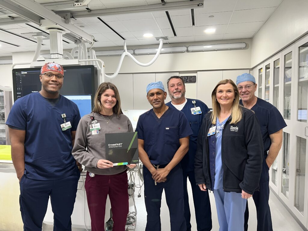 Six medical professionals, wearing scrubs and hospital badges, stand and pose together in a hospital room.