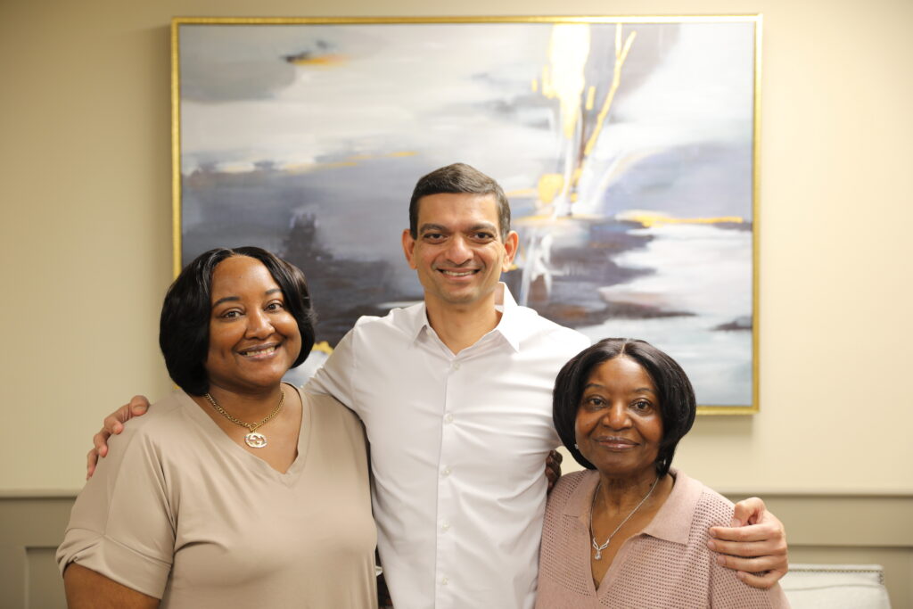 Three adults stand close together and smile at the camera in front of an abstract painting on a beige wall.