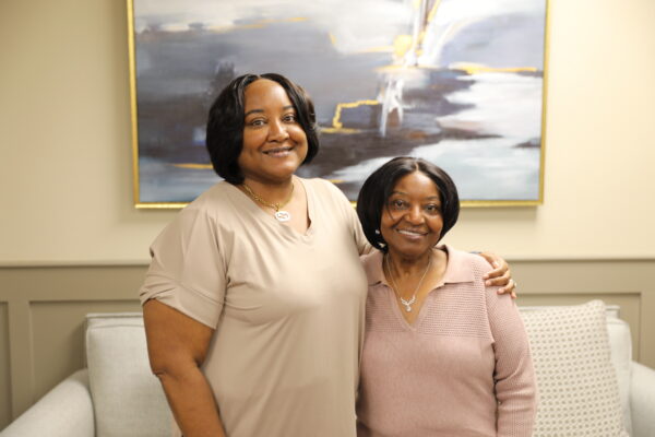 Two women stand side by side smiling in front of a couch and abstract painting, one with her arm around the other.