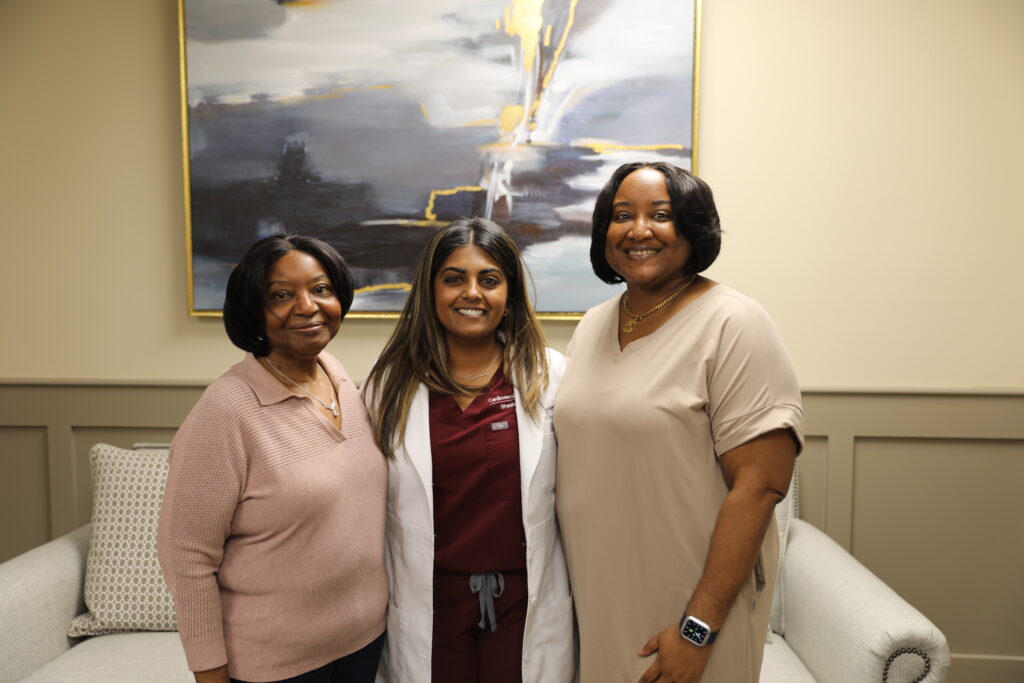 Three women stand together indoors, one wearing a white coat, in front of a sofa and abstract painting, all smiling at the camera.
