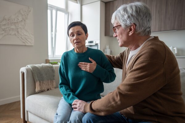 An older woman sits on a couch holding her chest, while an older man next to her touches her shoulder and knee, appearing concerned.
