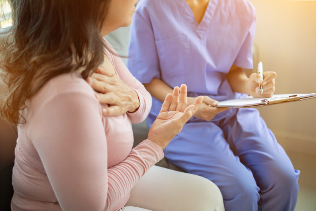 A woman holds her chest while talking to a healthcare worker in scrubs who is writing on a clipboard.