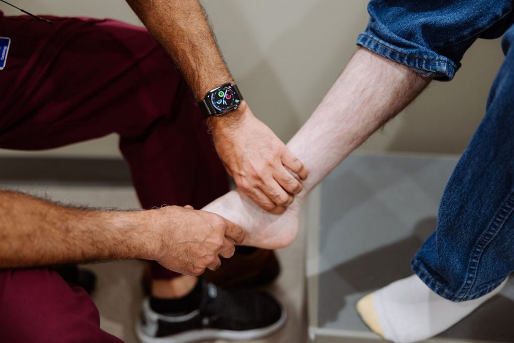 A nurse wearing scrubs examines another person’s bare ankle and foot while the patient sits with one sock off.
