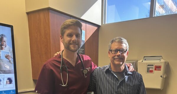 A healthcare worker in burgundy scrubs stands next to a smiling man in casual clothes inside a medical office exam room.