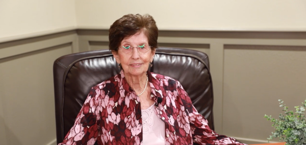An older woman with short brown hair sits on a dark leather chair, wearing glasses and a floral shirt, with a plant on the right.