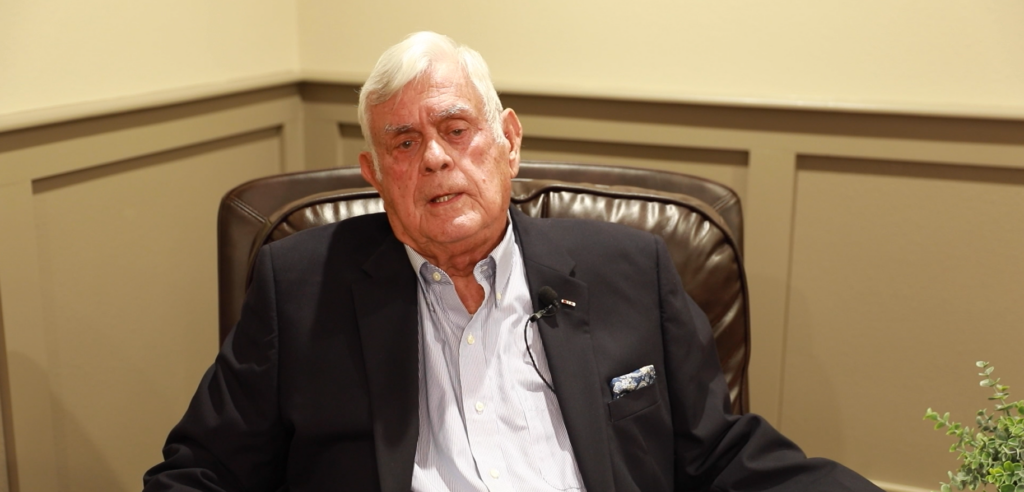 An older man in a dark blazer sits in a brown leather chair against a beige wall, looking forward with a neutral expression.